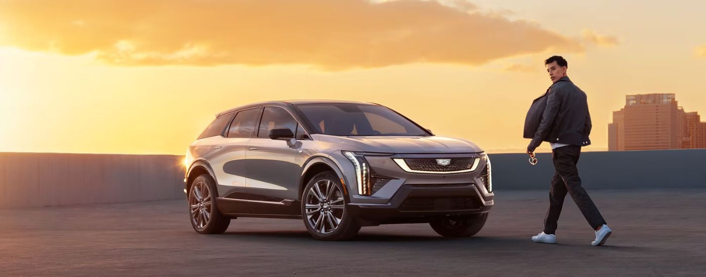 A man walking to a silver 2025 Cadillac OPTIQ parked at sunset.